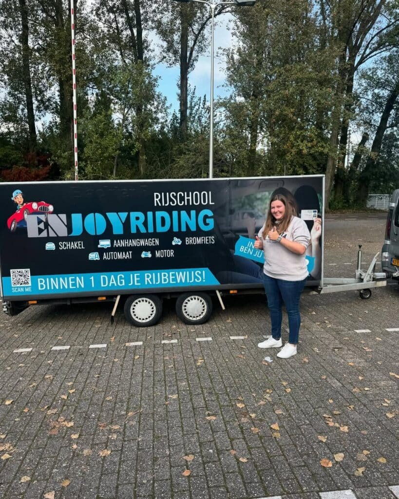 Geslaagde cursiste met rijbewijs voor rijschool trailer op parkeerplaats Geslaagde cursiste met rijbewijs voor rijschool trailer op parkeerplaats